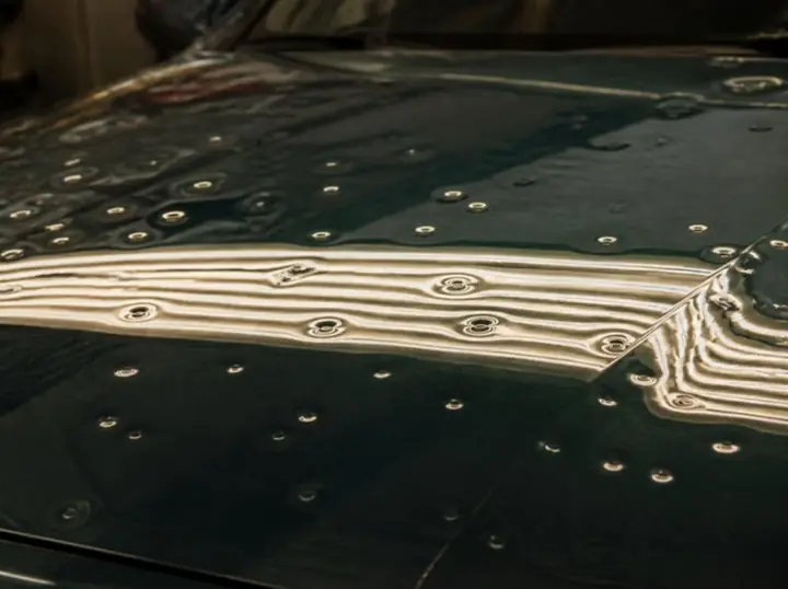Dark green hood covered in dozens of small hail dents under a striped inspection light