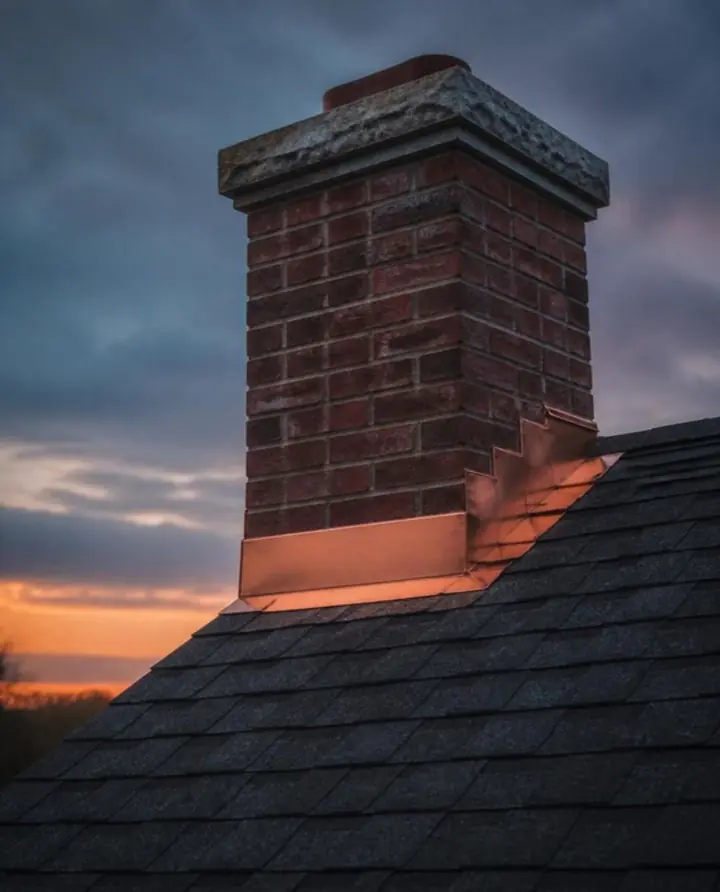 A brick chimney with fresh copper step flashing on a dark shingle roof at dusk
